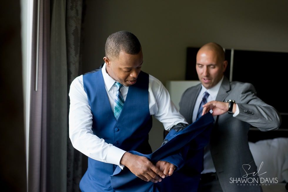 Two men in suits adjusting a blue vest indoors near a window