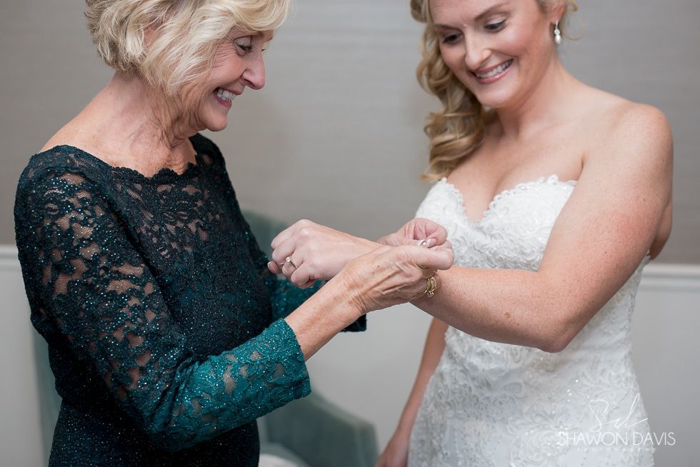 Older woman fastens a bride’s wedding dress strap as they smile together indoors