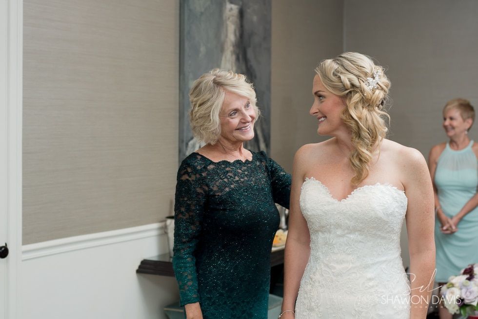 Bride in white dress smiling with an older woman in a dark green dress indoors.