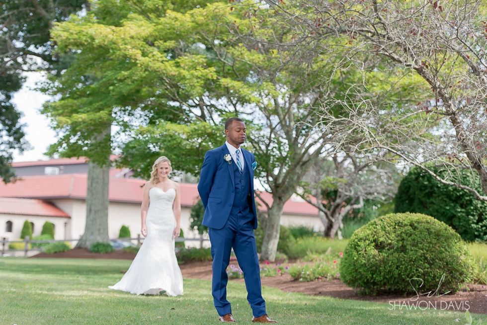 Two people in formal attire standing on a lawn with trees and a building in the background.