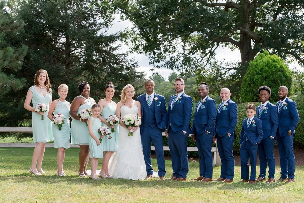 Wedding party posing outdoors on a lawn, with bridesmaids in mint dresses and groomsmen in blue suits.