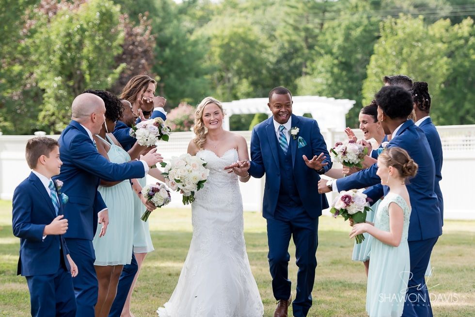 Wedding party cheers as bride and groom walk down an outdoor path, holding bouquets and smiling