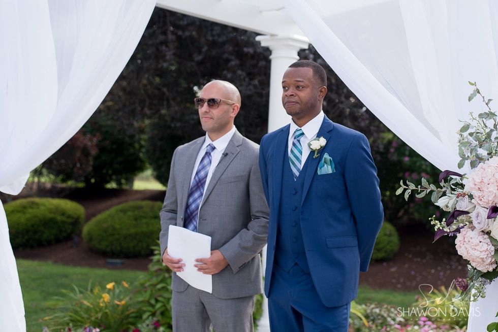 Two men in suits standing at an outdoor wedding arch with white drapes and flowers