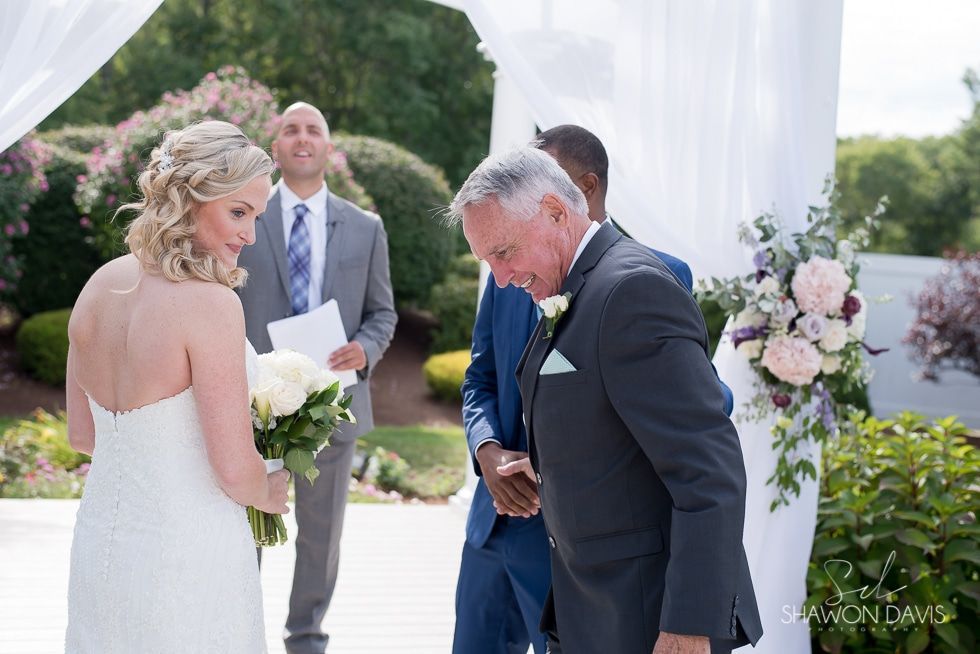 Bride in white dress faces groom in dark suit during outdoor wedding ceremony with floral arrangements.