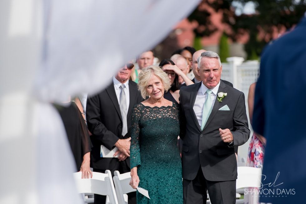 Couple walking arm in arm at an outdoor ceremony, woman in teal dress, man in dark suit.