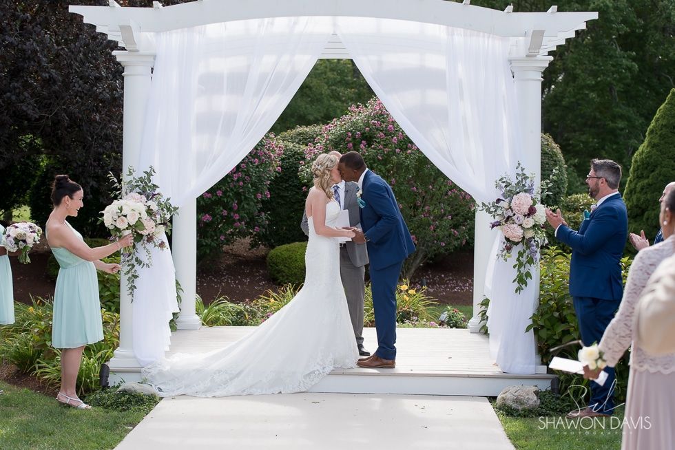 Wedding couple kissing under a white arbor, surrounded by floral arrangements and guests outdoors