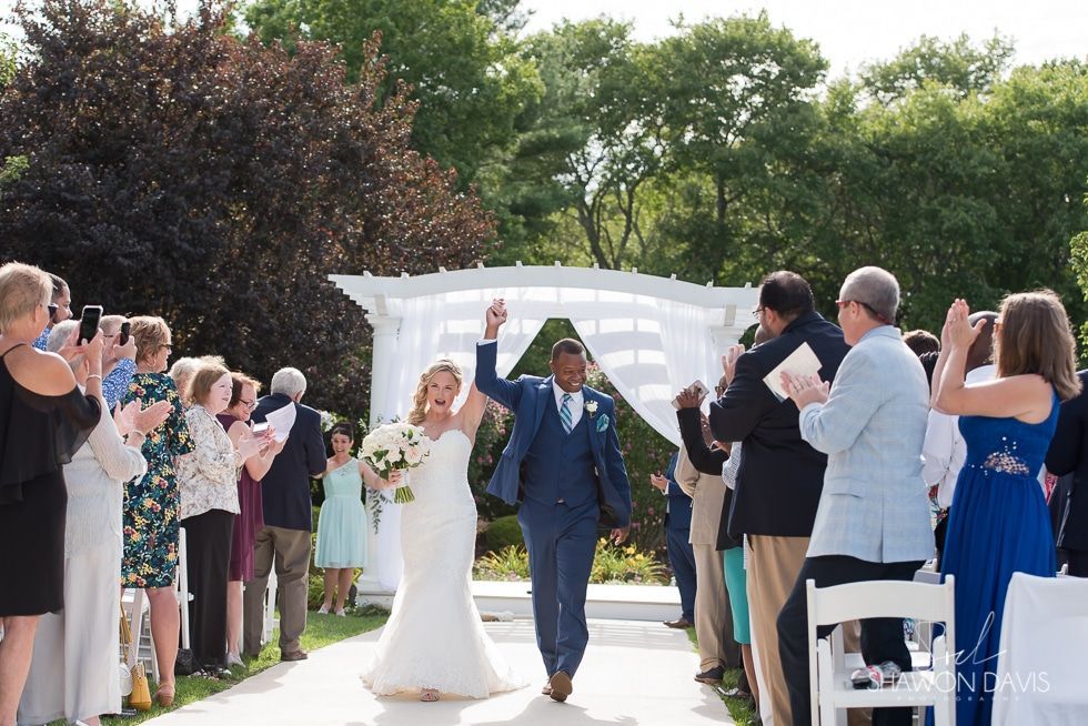 Bride and groom walking down an outdoor wedding aisle as guests applaud and take photos