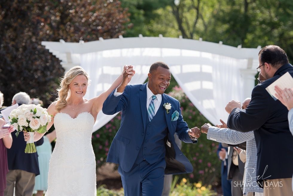 Newlyweds holding hands and walking down the aisle outdoors, surrounded by guests and flowers