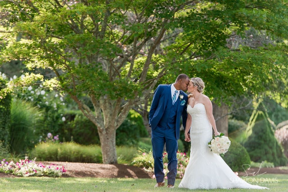 Bride and groom in blue suit and white gown sharing a kiss in a lush garden