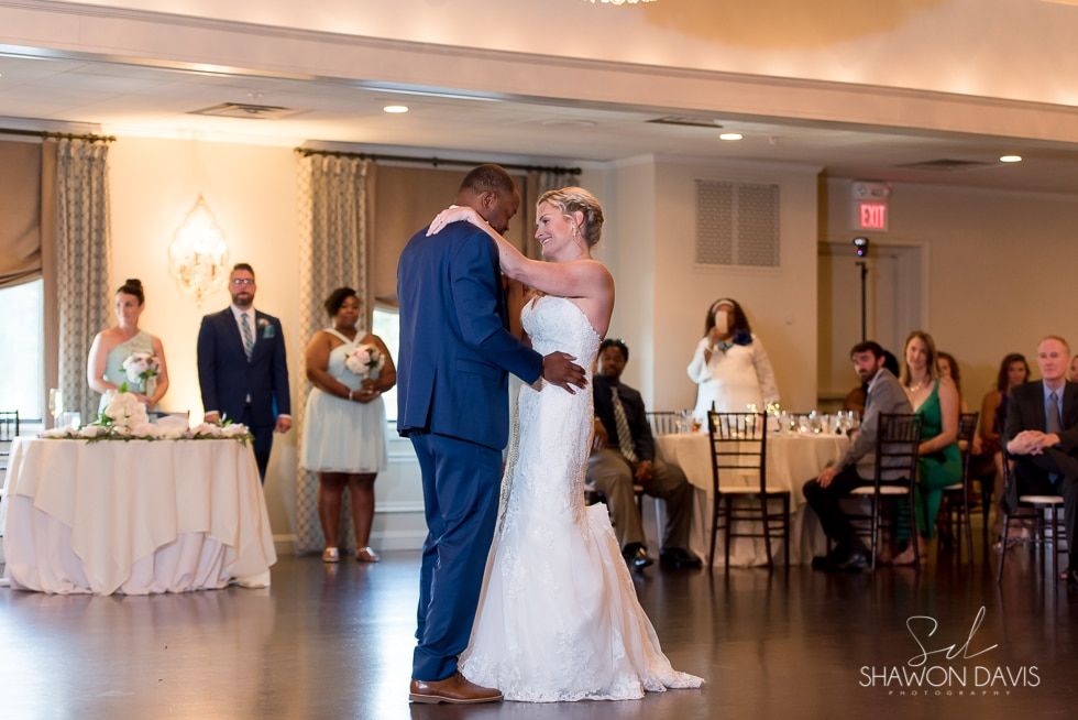 Bride and groom share a dance at a wedding reception, guests seated nearby.