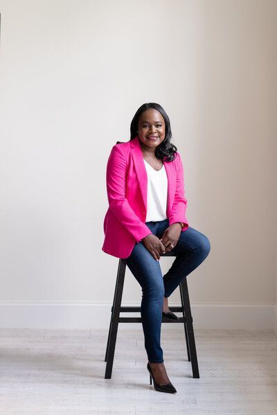 Woman seated on a stool in a pink blazer and jeans, smiling in a bright studio setting