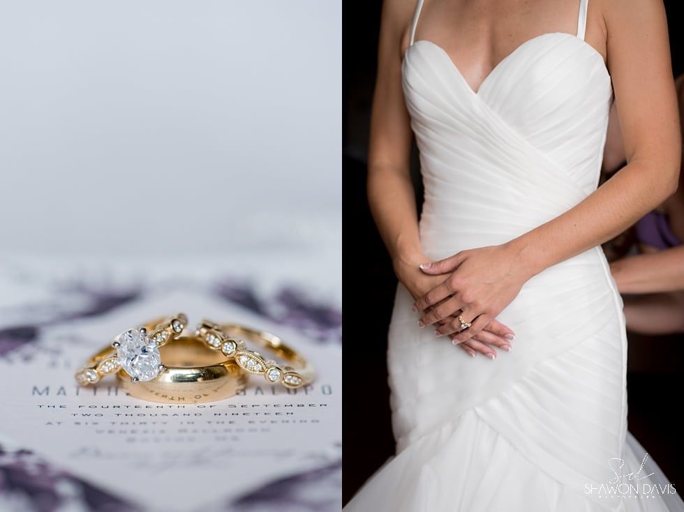 White wedding dress beside gold rings on a lace table, with the bride’s torso in the background