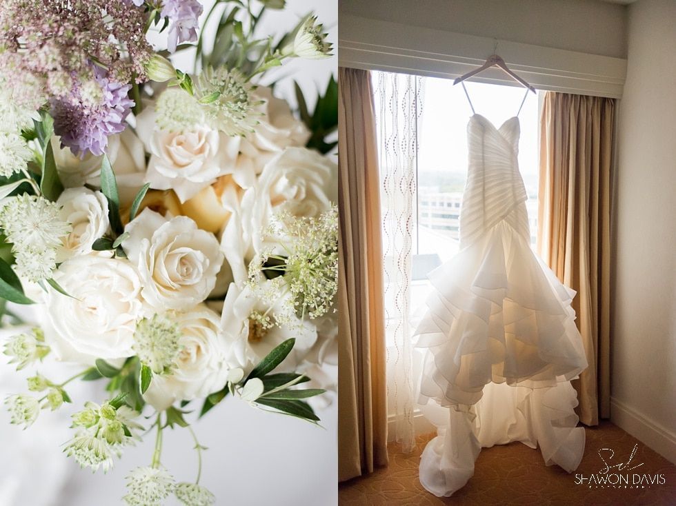 White wedding dress hanging by a bright window beside a bouquet of white and lavender flowers