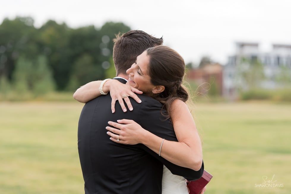 Two people embracing outdoors in a grassy field, with trees and buildings in the background.
