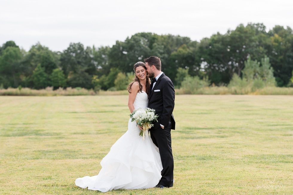 Bride and groom embracing in a field, holding a white bouquet, with trees in the background.