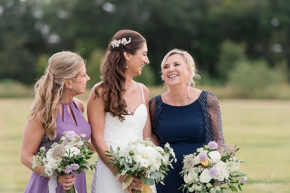 Three women in dresses holding bouquets outdoors, smiling and facing each other.