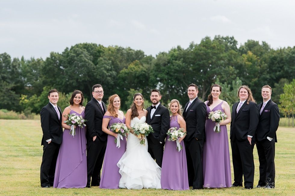 Wedding party posing in a field, with bridesmaids in lavender dresses and groomsmen in black suits.