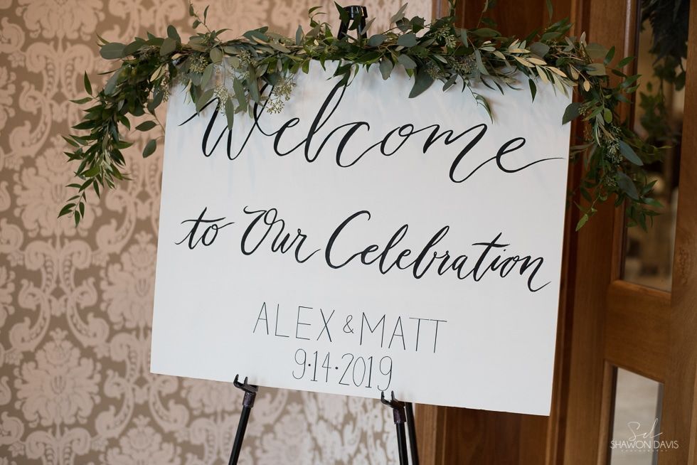Wedding welcome sign on easel with greenery, reading “Welcome to Our Celebration” and “Alex & Matt 9.14.19”
