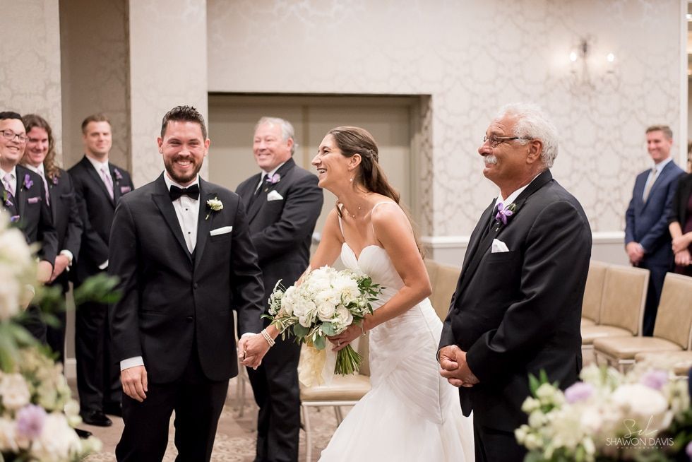 Bride and groom smiling during wedding ceremony, with guests standing nearby in a bright chapel.