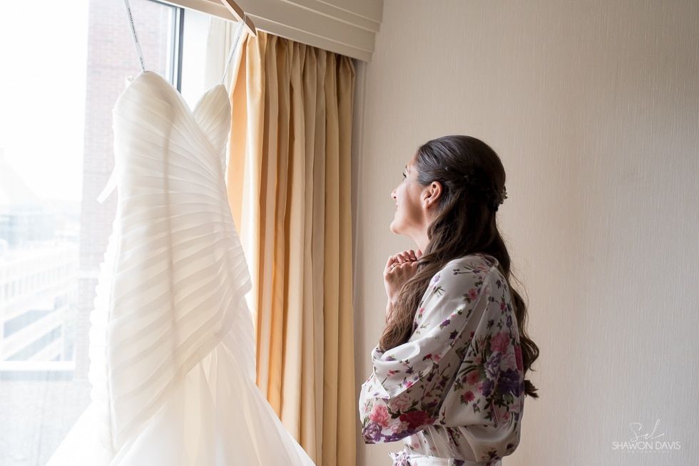 Woman in floral robe by a sunlit window, looking at a white wedding dress hanging nearby