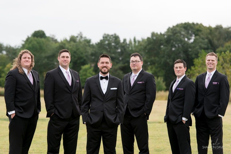 Six people in black suits standing outdoors on a grassy field with trees in the background