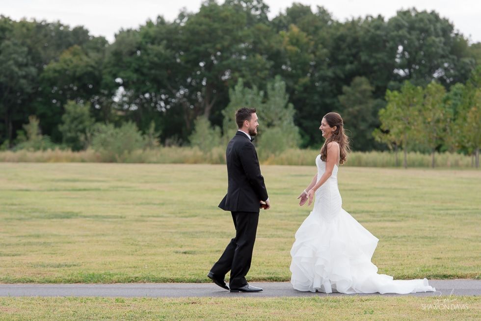 Bride and groom facing each other on a grassy field, with trees in the background.