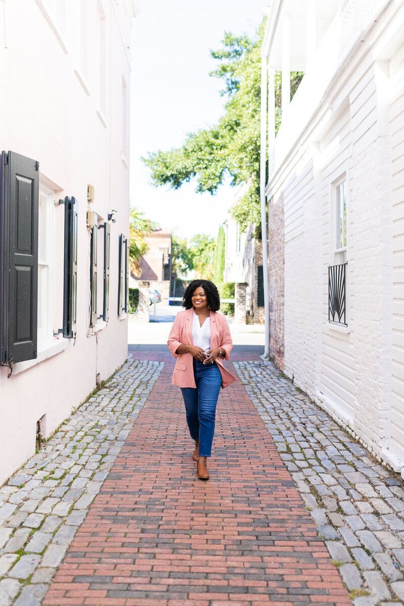 Person walking down a brick alley between white buildings, holding a pink jacket and a bag.