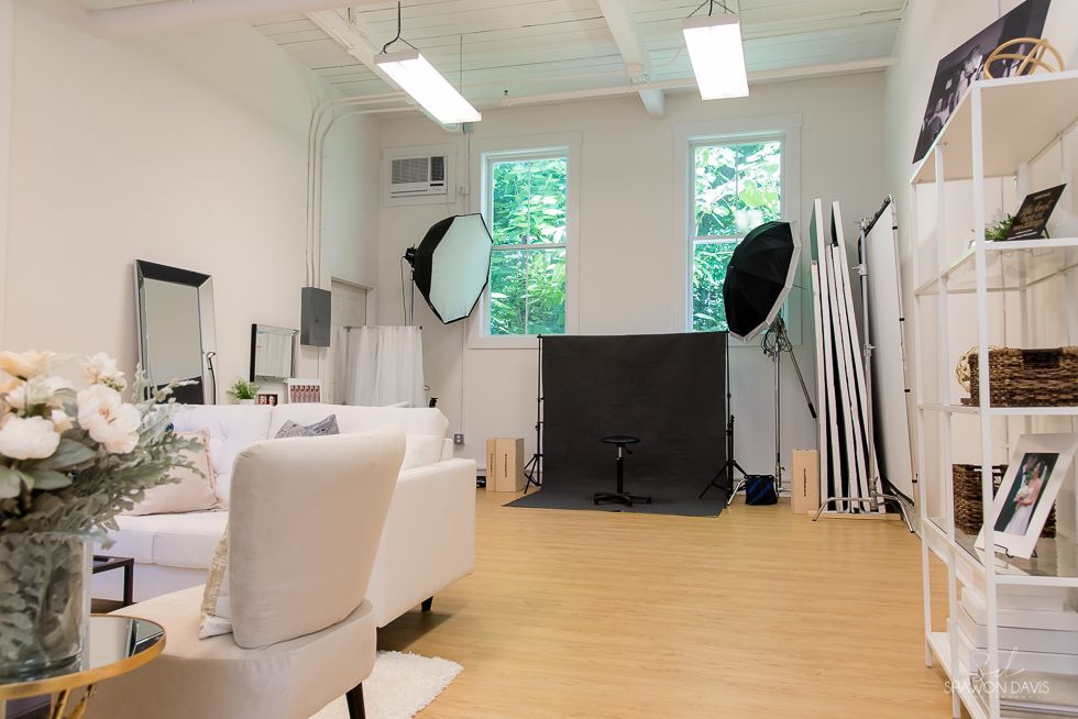 Bright studio with white sofa, large windows, lighting equipment, and stacked photo backdrops on the right.