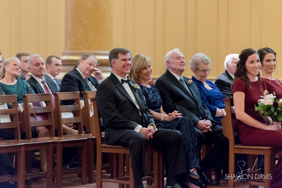 Guests seated in a church, including a man in a black suit and women holding bouquets.