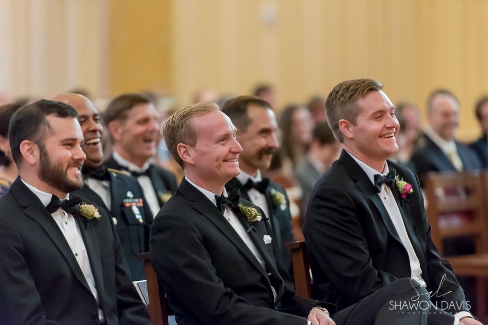 Three men in dark suits smile at a formal indoor ceremony, seated in a row.