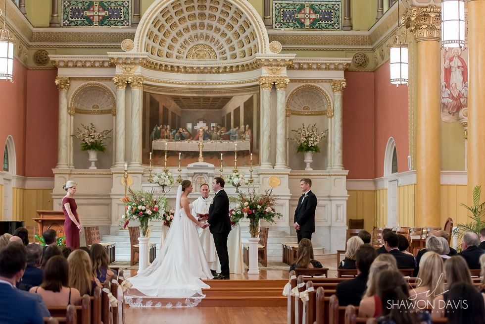 Bride and groom at altar in ornate church, with bridesmaids and guests seated nearby