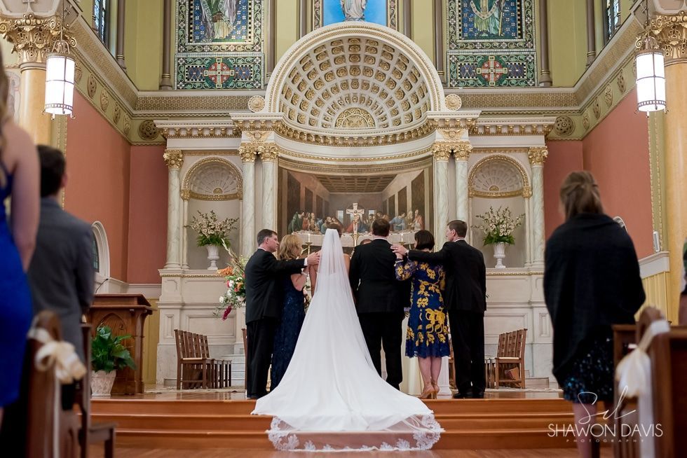 Bride and groom at the altar in a decorated church, seen from behind, with guests standing nearby