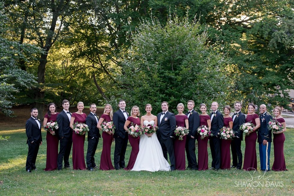 Wedding party posing outdoors in burgundy and black attire, with bride in white holding bouquet.