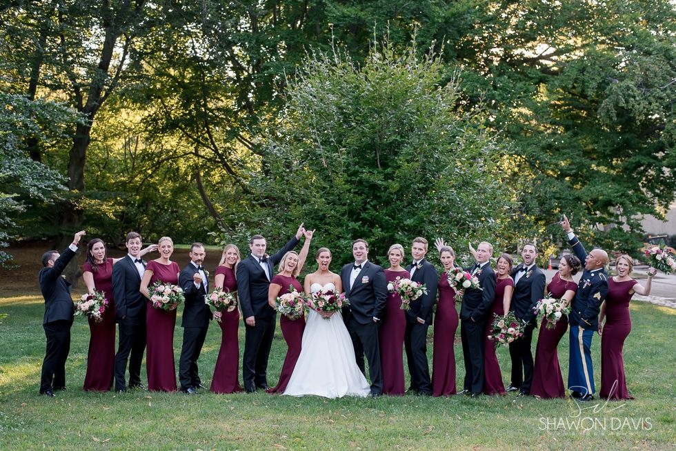 Wedding party posing outdoors, bride in white centered among attendants in burgundy dresses holding bouquets