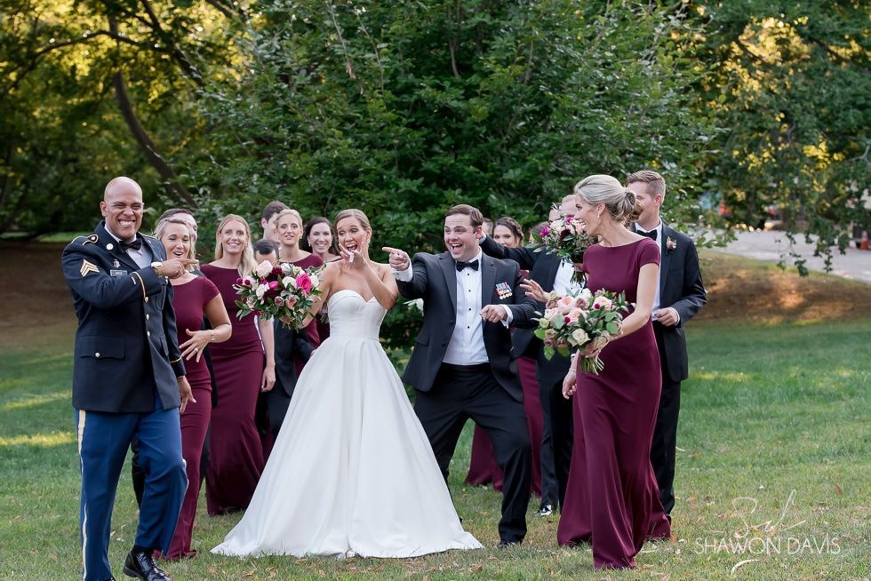 Wedding party posing outdoors, bride in white and attendants in burgundy holding champagne glasses.