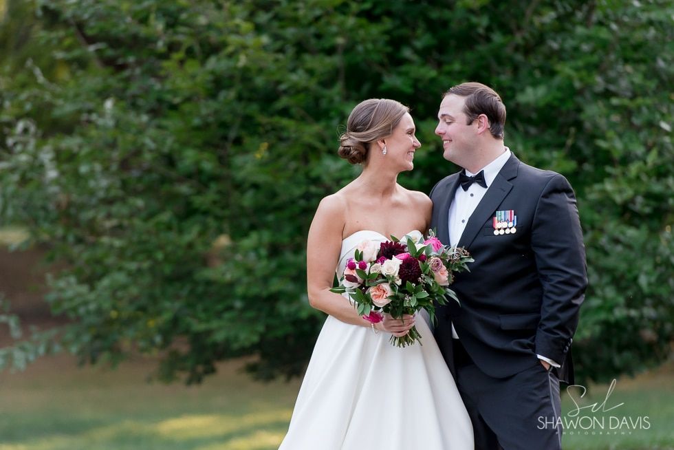 Bride and groom smiling at each other outdoors, bride holding a bouquet in a white gown