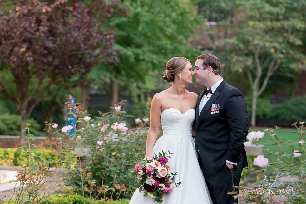 Bride and groom standing in a garden, smiling and holding a bouquet at their wedding.