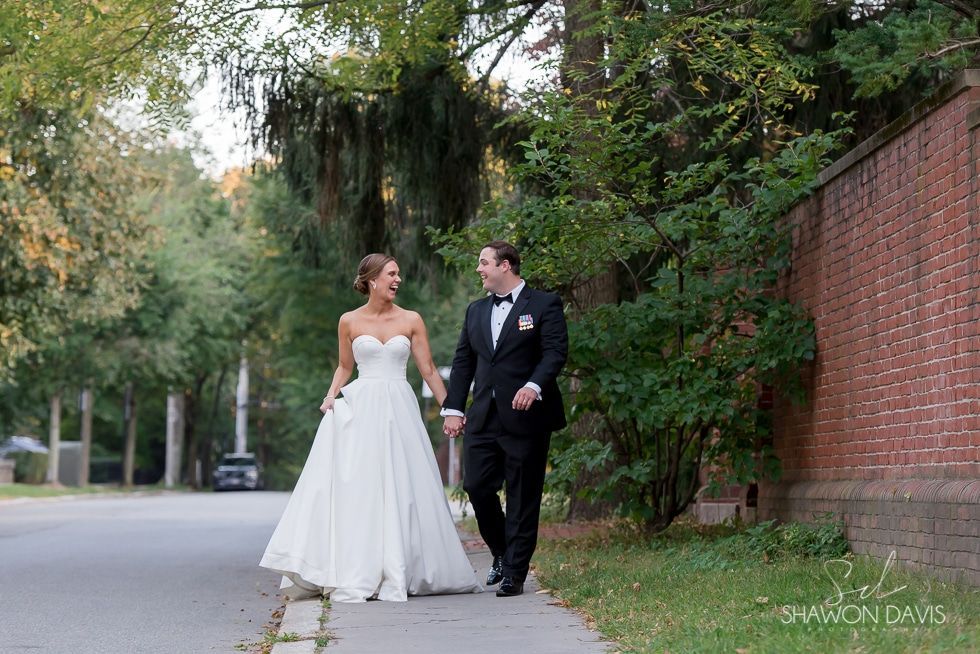 Bride and groom walking together on a leafy sidewalk beside a brick wall
