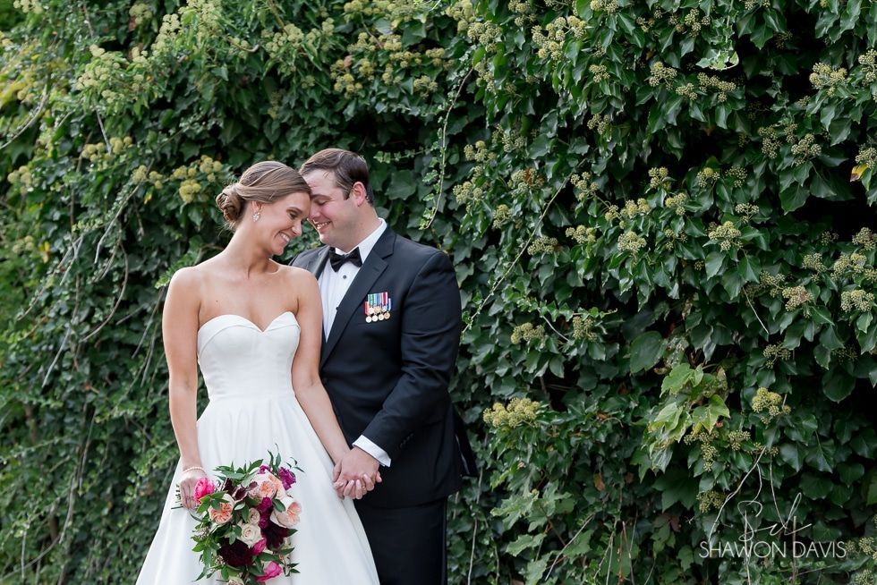 Bride and groom in wedding attire standing close together before a leafy green backdrop