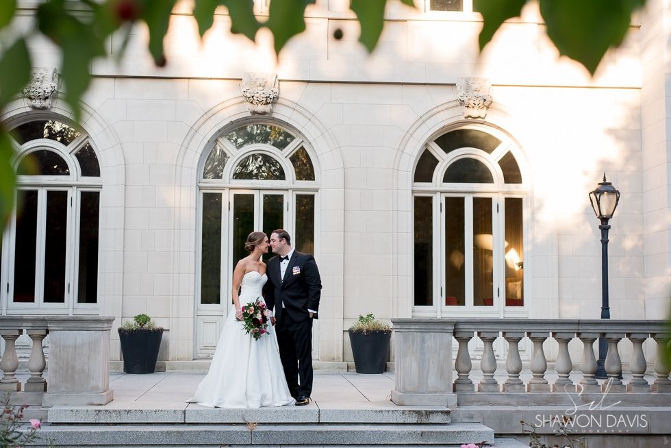 Bride and groom posing in front of elegant white arched windows and stone balustrade at sunset