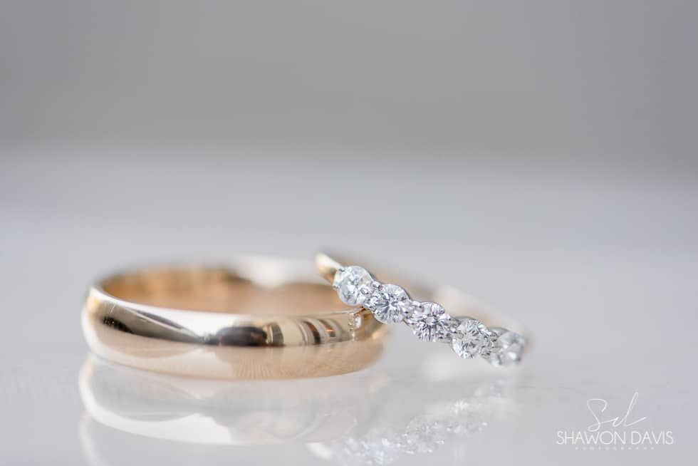 Gold wedding band beside a diamond engagement ring on a white reflective surface