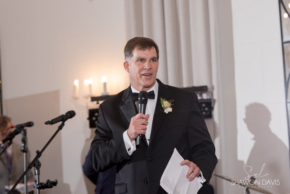 Man in a black tuxedo speaking into a microphone at an indoor event, holding papers.