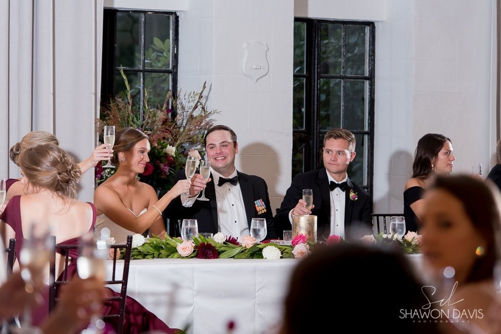 Guests raise glasses at a formal banquet table in a bright white reception hall.