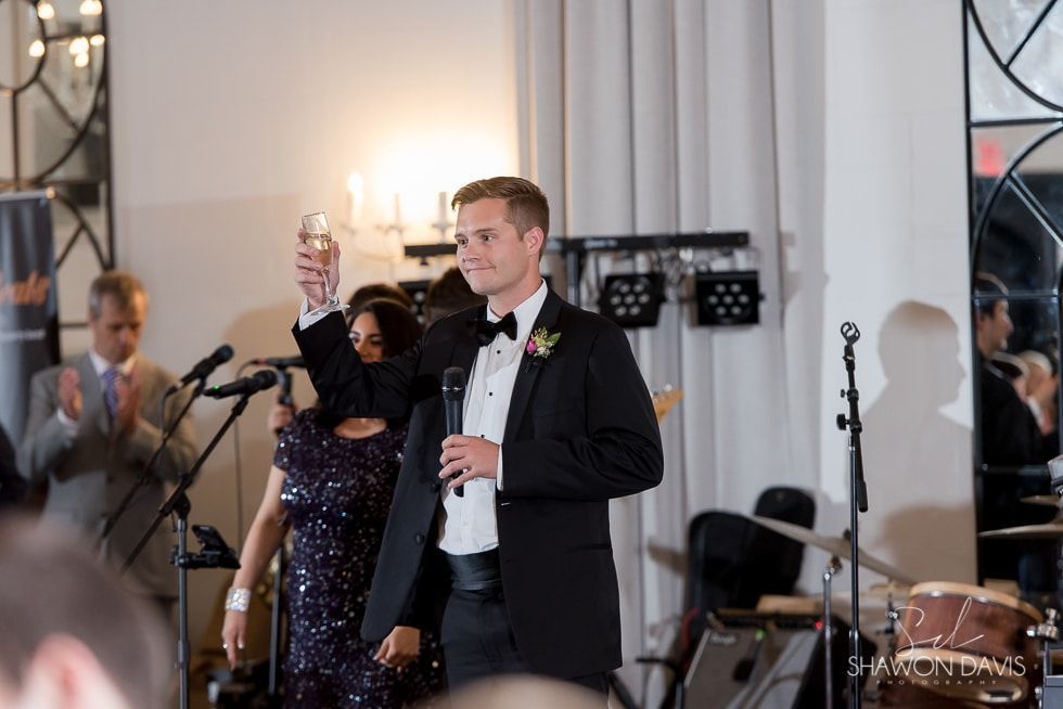 Man in tuxedo toasting at a formal indoor event with musicians in the background