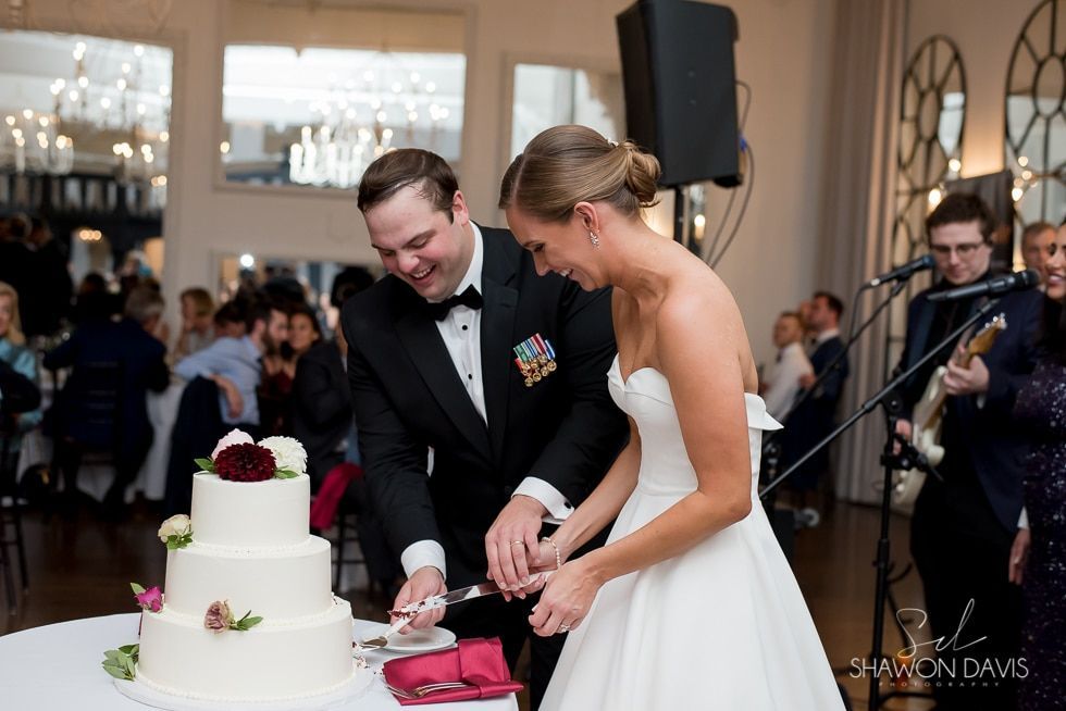 Bride and groom cut their wedding cake at a reception hall, smiling beside a three-tier white cake.