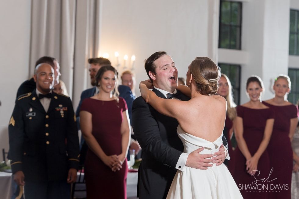 Bride and groom embrace during a wedding ceremony, with officiants and bridesmaids in the background.