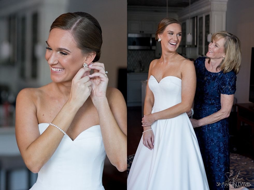 Bride in white strapless gown smiles while adjusting an earring as another woman helps her in a room.