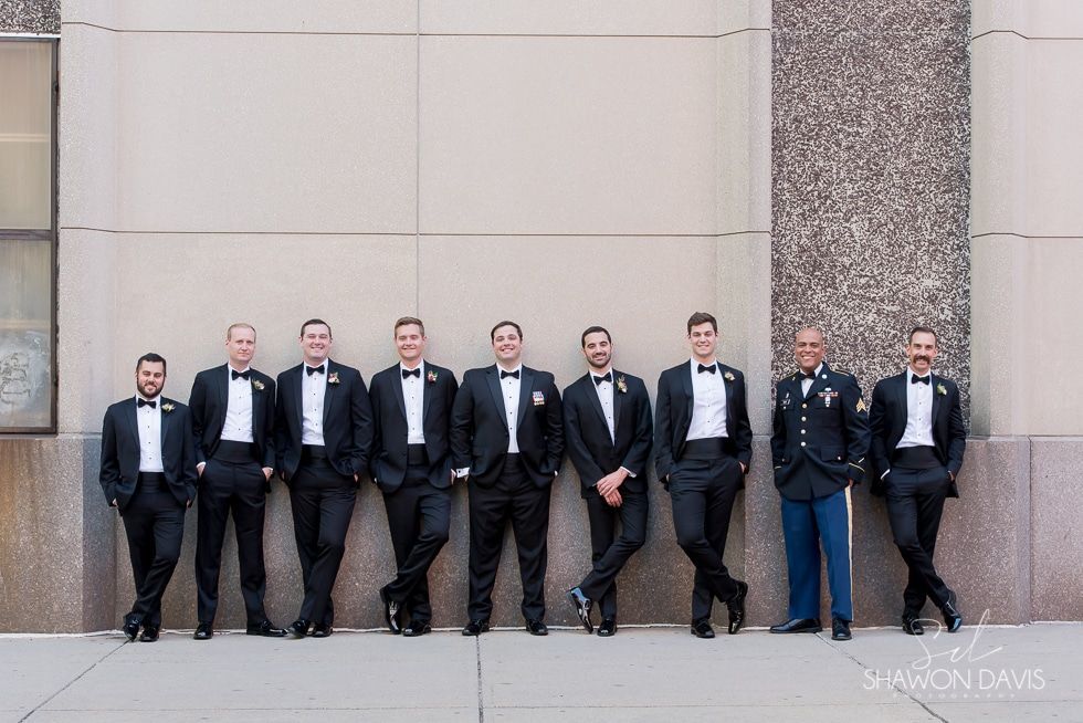 Nine people in dark suits stand lined up against a stone wall outside a building.