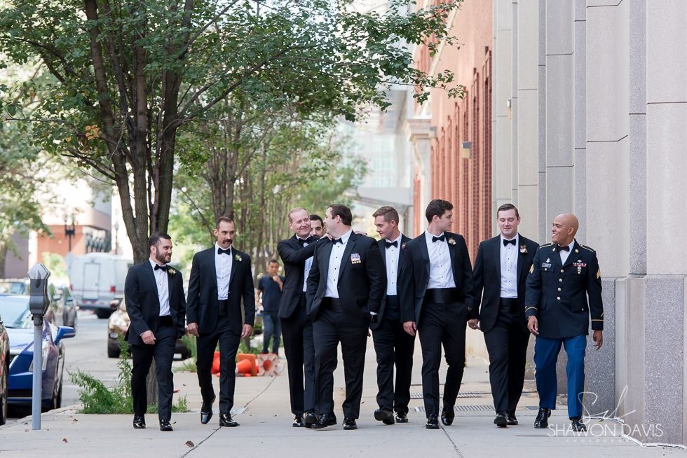 Six men in black suits walk together on a city sidewalk beside a building.