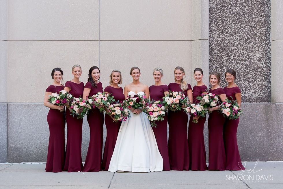 Nine women in burgundy dresses and one in white hold bouquets against a stone wall.